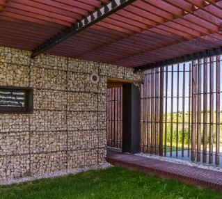Voladizo de madera sobre la entrada de un edificio con muro de gaviones
